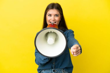 Young caucasian woman isolated on yellow background shouting through a megaphone to announce something while pointing to the front