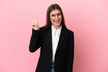 Genç beyaz kadın pembe arka planda izole edilmiş gülümsüyor ve zafer işareti gösteriyor.