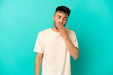 Kafkasyalı genç adam mavi arka planda izole edilmiş esniyor ve geniş açık ağzını eliyle kapatıyor.