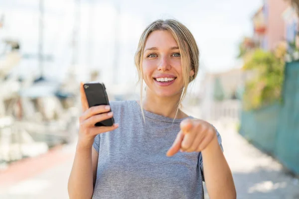 Young Blonde Woman Using Mobile Phone Outdoors Points Finger You — Stock Photo, Image