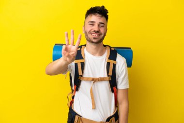 Sarı arka planda izole edilmiş büyük sırt çantalı beyaz bir genç adam mutlu ve parmaklarıyla üçe kadar sayıyor.