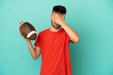 Genç beyaz adam rugby oynuyor. Mavi arka planda izole edilmiş. Gözlerini elleriyle kapatıyor. Bir şey görmek istemiyorum.