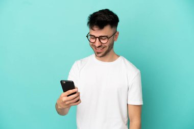 Mavi arka planda izole edilmiş genç beyaz adam cep telefonuyla bir mesaj ya da e-posta gönderiyor