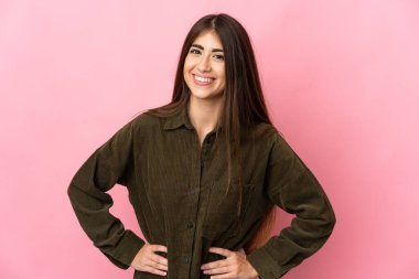 Pembe arka planda izole edilmiş genç beyaz kadın kolları kalçasında ve gülümsüyor.