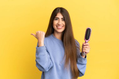 Young caucasian woman holding hairbrush isolated on blue background pointing to the side to present a product