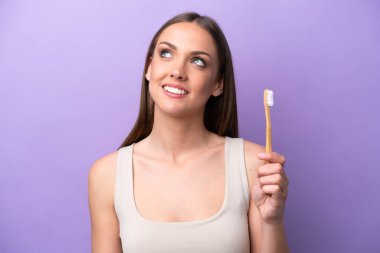 Mor arka planda dişlerini fırçalayan beyaz bir kadın gülümserken yukarı bakıyor.