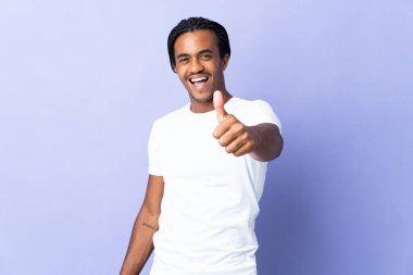 Örgülü genç Afrikalı Amerikalı adam mor arka planda izole edildi. Başparmakları havada çünkü iyi bir şey oldu.