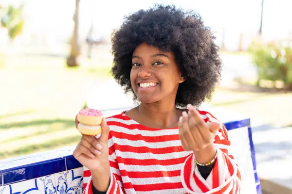 African American girl holding a donut at outdoors inviting to come with hand. Happy that you came