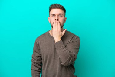 Genç beyaz adam mavi arka planda izole edilmiş, ağzı elle kapatılmış.