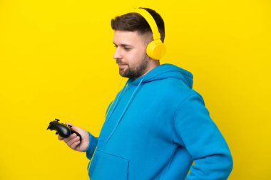 Bir video oyunu kontrolörüyle oynayan genç beyaz adam sarı arka planda izole edilmiş ve sırt ağrısı çekiyor.
