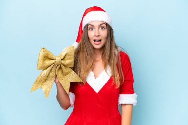 Anne Noel gibi giyinmiş genç beyaz kadın elinde Noel yayı ile mavi arka planda izole edilmiş, şaşırmış ve şok olmuş bir yüz ifadesiyle.