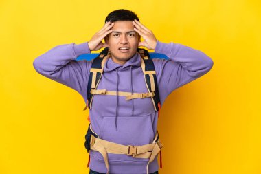 Sarı arka planda büyük bir sırt çantası olan genç dağcı, sürpriz bir ifadeyle izole edilmiş.