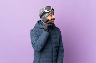 İzole edilmiş mor arka planda snowboard gözlüklü kayakçı cep telefonuyla sohbet ediyor.