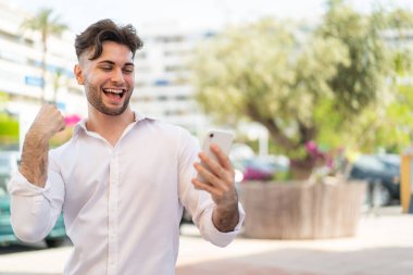 Dışarıda cep telefonu kullanan yakışıklı genç bir adam zaferi kutluyor.