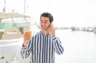 Genç yakışıklı adam cep telefonu kullanıyor ve elinde mutlu bir ifadeyle kahve tutuyor.