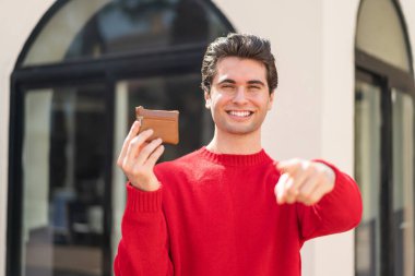 Açık havada cüzdan tutan genç yakışıklı bir adam kendinden emin bir ifadeyle seni işaret ediyor.