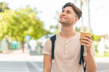 Genç yakışıklı bir adam açık havada kahve alıp gülümseyerek yukarı bakıyor.