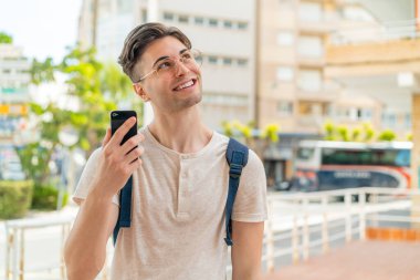 Dışarıda cep telefonu kullanan genç yakışıklı adam gülümserken yukarı bakıyor.