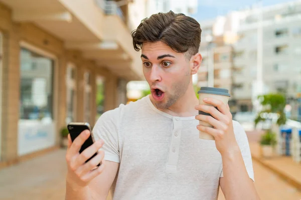 Mladý Pohledný Muž Pomocí Mobilního Telefonu Drží Kávu Překvapeným Výrazem — Stock fotografie