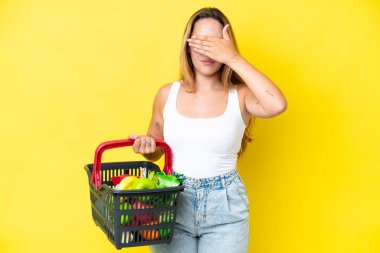 Young caucasian woman holding a shopping basket full of food isolated on yellow background covering eyes by hands. Do not want to see something