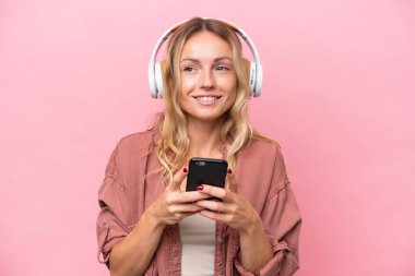 Pembe arka planda izole edilmiş genç bir Rus kadın cep telefonuyla müzik dinliyor ve ön tarafa bakıyor.