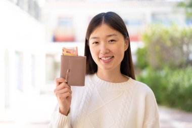 Güzel bir Çinli kadın elinde cüzdanla dışarıda mutlu bir ifadeyle bekliyor.