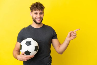 Yakışıklı genç futbolcu, sarı arka planda izole edilmiş, parmağıyla kenarı işaret ediyor.