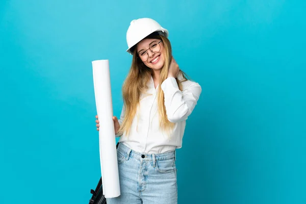 Young architect woman with helmet and holding blueprints isolated on blue background laughing