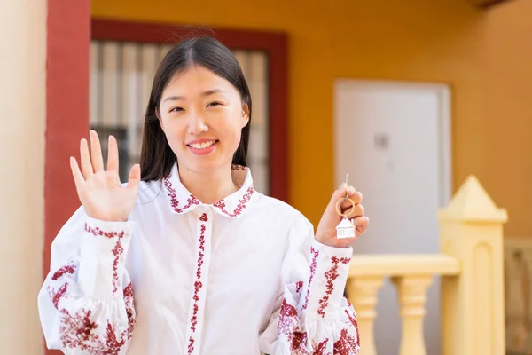 Young Chinese woman holding home keys at outdoors saluting with hand with happy expression
