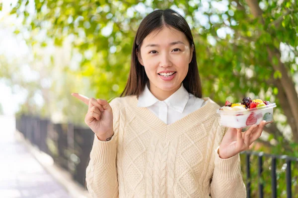 Young Chinese Woman Holding Bowl Fruit Outdoors Surprised Pointing Finger — Stock Photo, Image