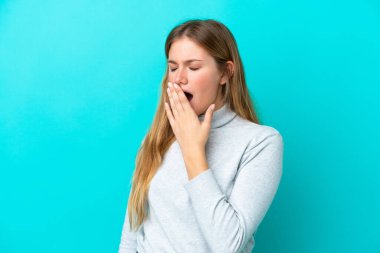 Genç sarışın kadın mavi arka planda izole edilmiş esniyor ve geniş bir ağzı eliyle kapatıyor.