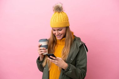 Kışlık ceket giyen genç sarışın kadın pembe arka planda izole edilmiş. Elinde kahve ve cep telefonu var.