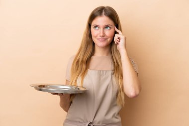 Bej arka planda saklanmış tepsisi olan genç sarışın garson kadının şüpheleri ve düşünceleri var.