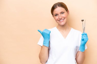 Beyaz dişçi kadının elinde bej arka planda izole edilmiş aletler var. Ürünü sunmak için yan tarafı işaret ediyor.