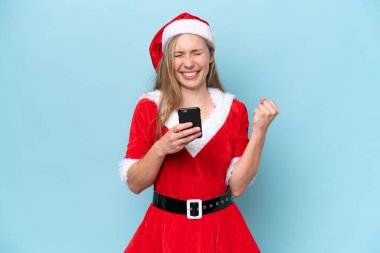 Anne Noel Baba gibi giyinmiş genç sarışın kadın mavi arka planda izole edilmiş ve telefon zafer pozisyonunda.