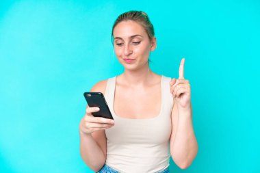 Genç beyaz kadın mavi arka planda cep telefonu ve parmak kaldırarak izole edilmiş.