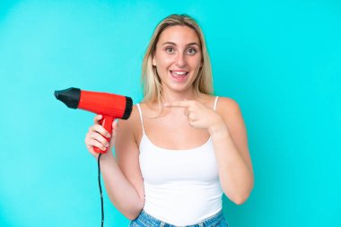 Kafkasyalı genç bir kadın elinde saç kurutma makinesi tutuyor mavi arka planda izole edilmiş ve onu işaret ediyor.