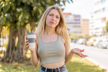 Sarışın bir kadın açık havada kahve içerken omuzlarını kaldırırken şüpheyle hareket ediyor.