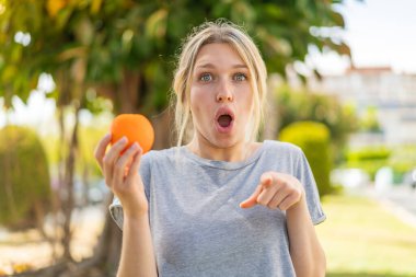 Açık havada portakal tutan genç sarışın kadın şaşırmış ve ön tarafı işaret ediyor.