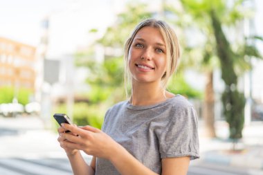 Dışarıdaki genç sarışın kadın cep telefonuyla mesaj ya da e-posta yolluyor.