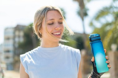 Açık havada bir şişe suyla spor yapan genç sarışın kadın.