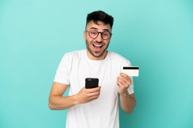 Kafkasyalı genç adam mavi arka planda izole edilmiş cep telefonuyla alışveriş yapıyor ve elinde şaşırmış bir ifadeyle bir kredi kartı tutuyor.