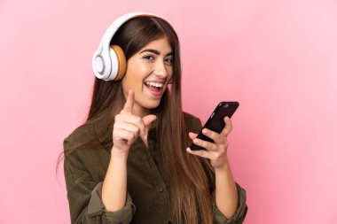 Pembe arka planda izole edilmiş genç beyaz bir kadın cep telefonuyla müzik dinliyor ve şarkı söylüyor.