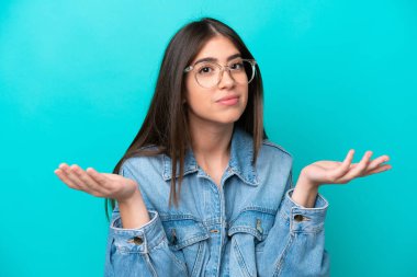 Genç ve güzel bir kadın şaşkın ve şaşkın hissediyor, şüpheci ve ağırlıklı ya da farklı seçenekleri seçiyor mavi duvara karşı komik bir ifadeyle