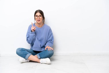 Beyaz arka planda izole bir şekilde yerde oturan genç beyaz bir kadın, bir fikrin işaret ettiğini düşünüyor.