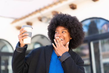 Afro-Amerikalı bir kız elinde sürpriz ve şok edici bir yüz ifadesiyle açık havada kahve tutuyor.