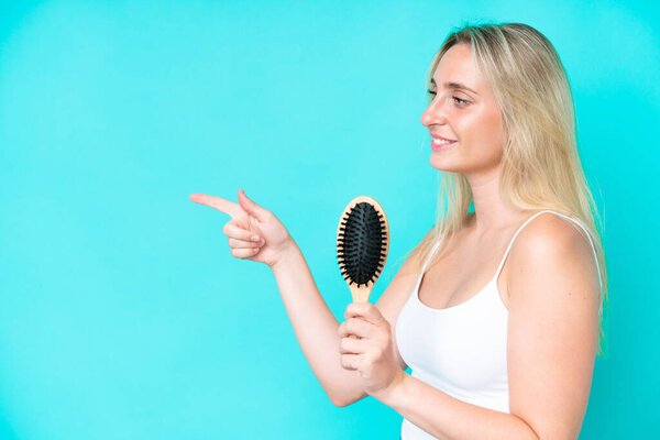 Young caucasian woman with hair comb isolated on blue background pointing to the side to present a product