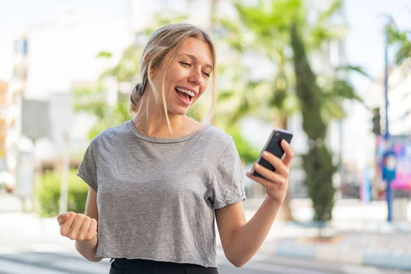 Young Blonde Woman Outdoors Using Mobile Phone Doing Victory Gesture — Stock Photo, Image