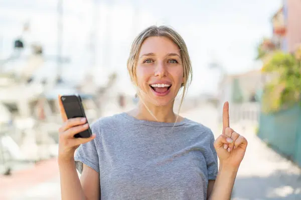Young Blonde Woman Using Mobile Phone Outdoors Pointing Great Idea — Stock Photo, Image