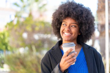 Afro-Amerikalı bir kız açık havada elinde neşeli bir ifadeyle kahve tutuyor.